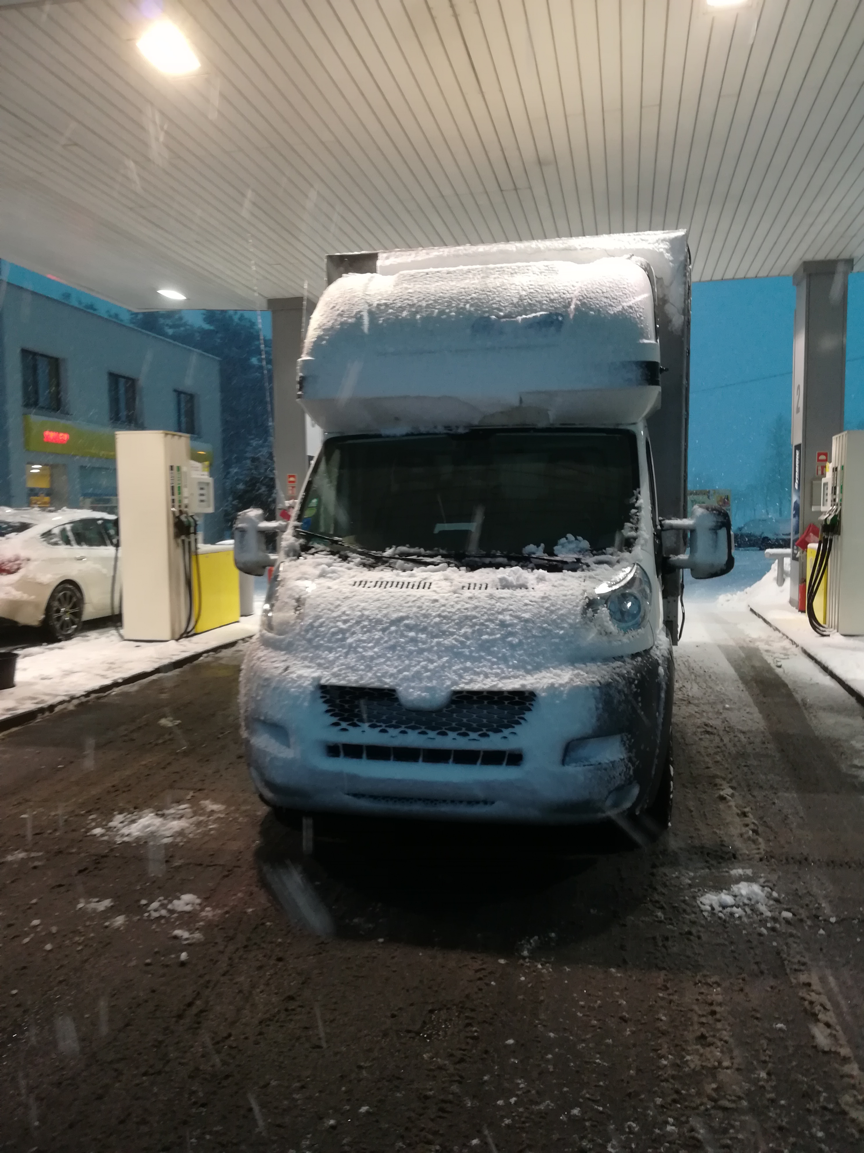 Przód białego busa zaparkowanego na stacji benzynowej, obficie pokrytego śniegiem podczas opadów śniegu.