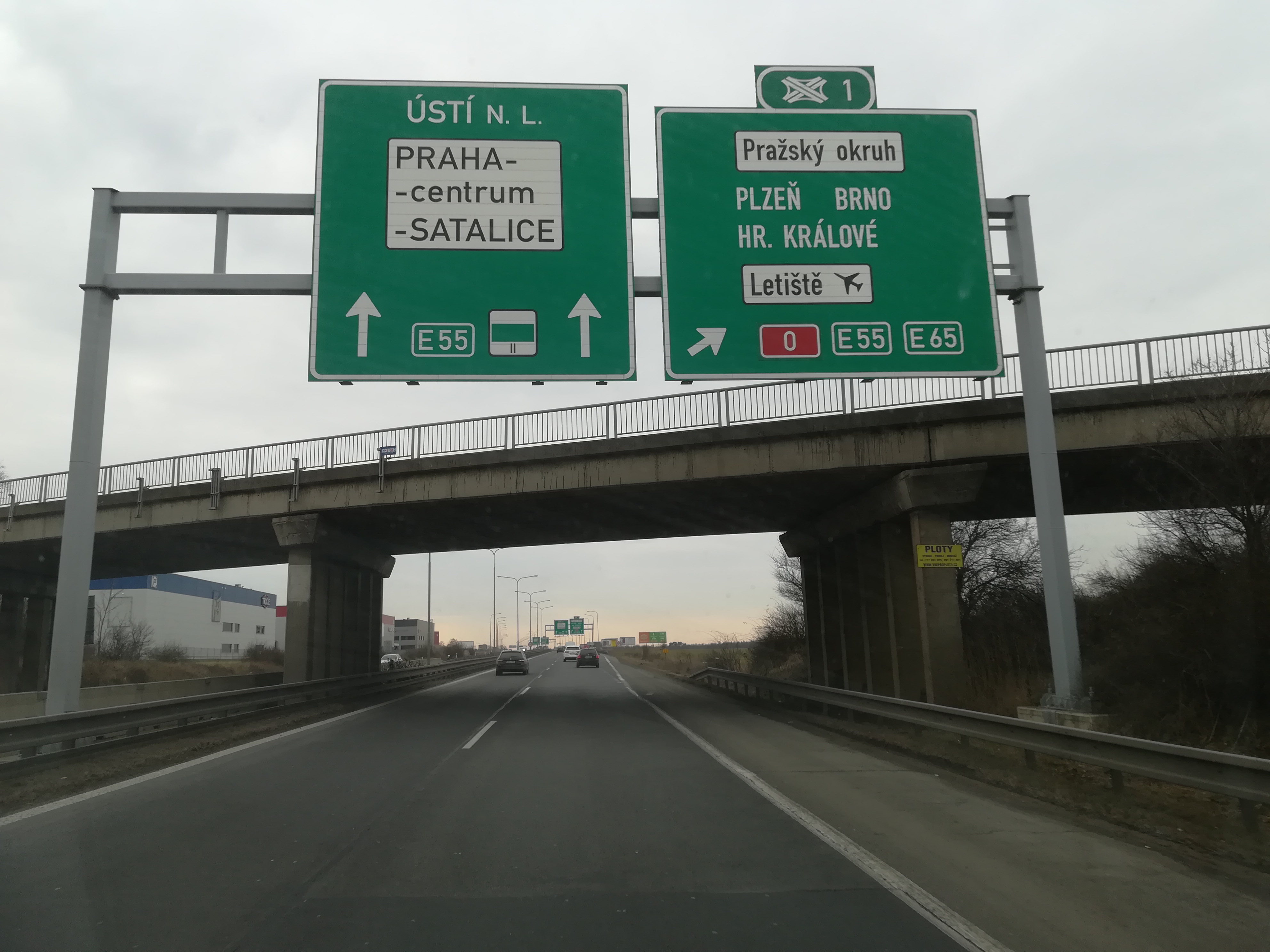 Widok z perspektywy kierowcy na autostradę z oznakowaniem kierunkowym do Pragi, Pilzna, Brna i Hradca Králové. Nad autostradą wiadukt z szarymi barierami.