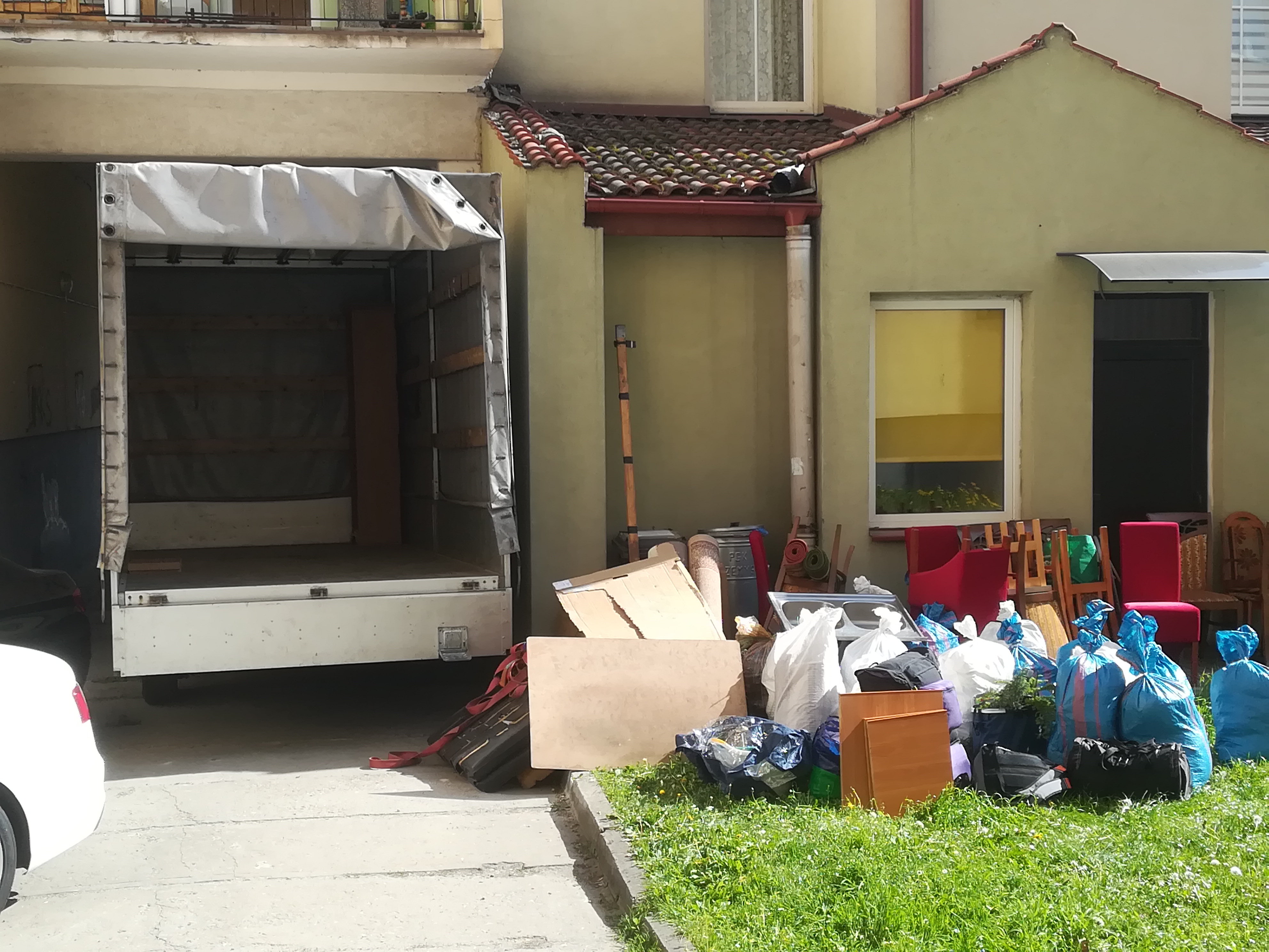 Otwarta paka busa na tle budynku z zebranymi rzeczami do transportu: worki, meble, kartony, dywan.