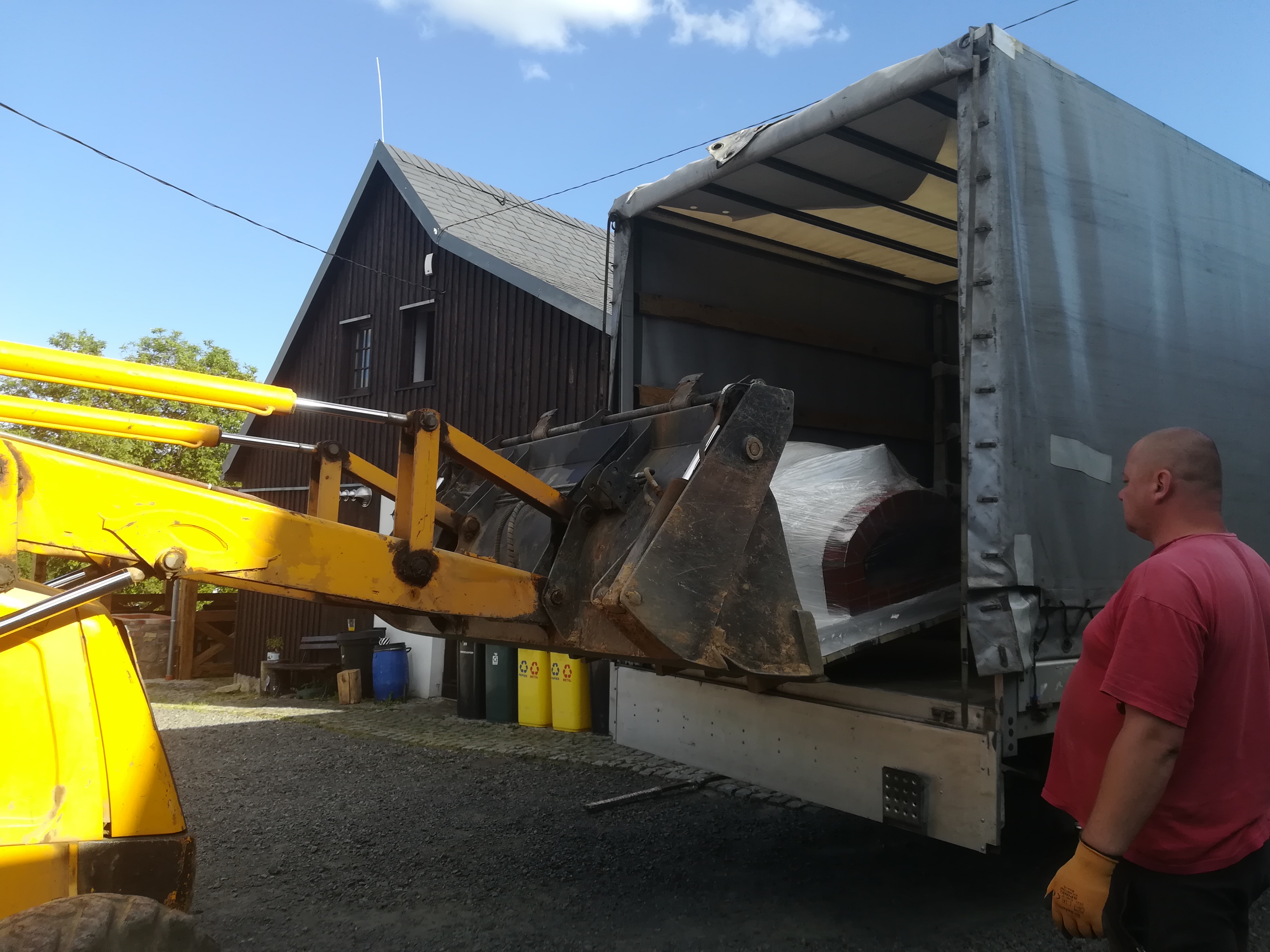 Żółta ładowarka teleskopowa z łyżką umieszcza zapakowany w folię element w otwartej naczepie ciężarówki; w tle budynek z ciemnego drewna i pojemniki na śmieci.