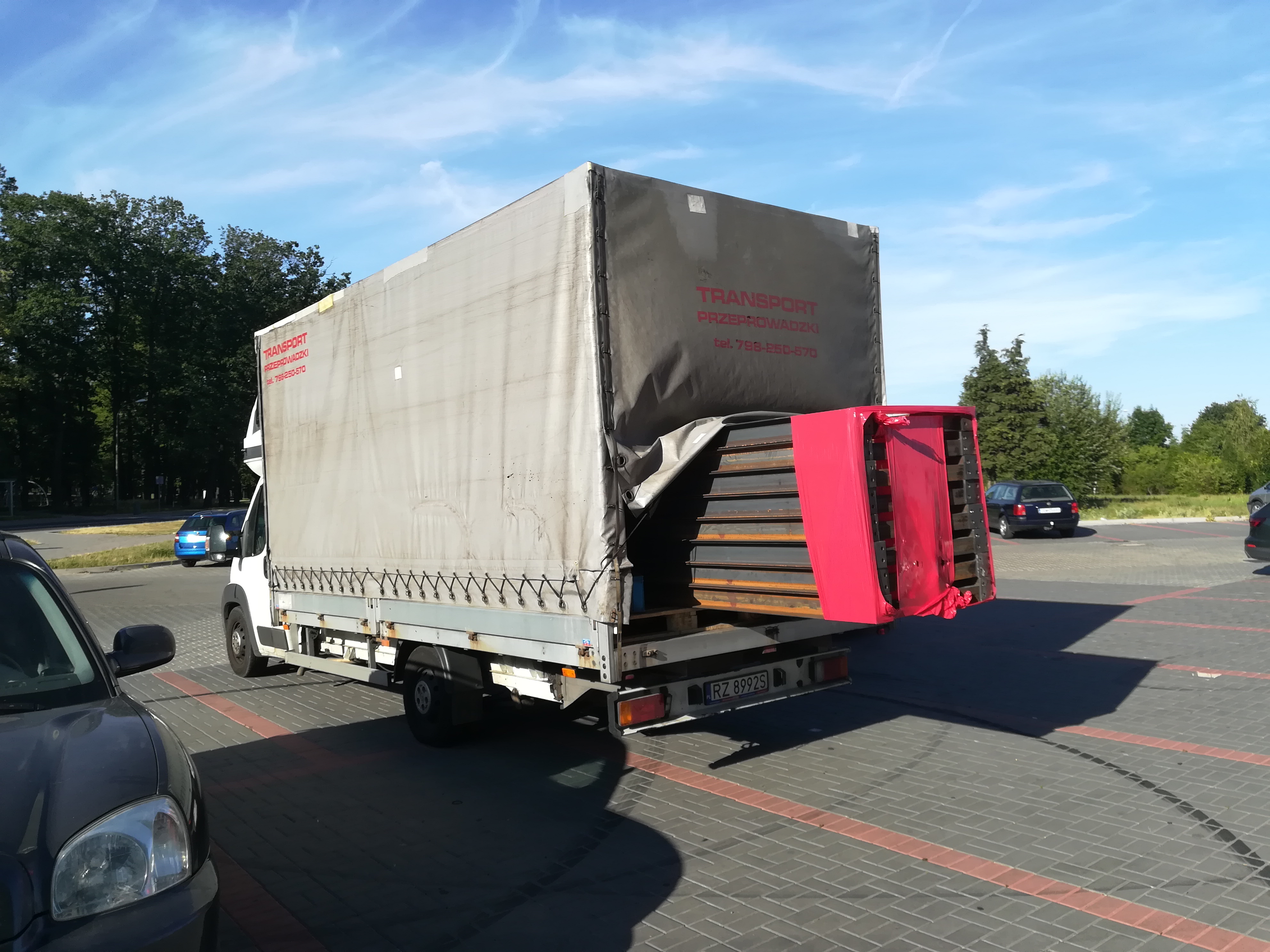 Ciężarówka z plandeką, z otwartą tylną klapą, załadowana elementami metalowymi w czerwonym opakowaniu, zaparkowana na parkingu w słoneczny dzień.