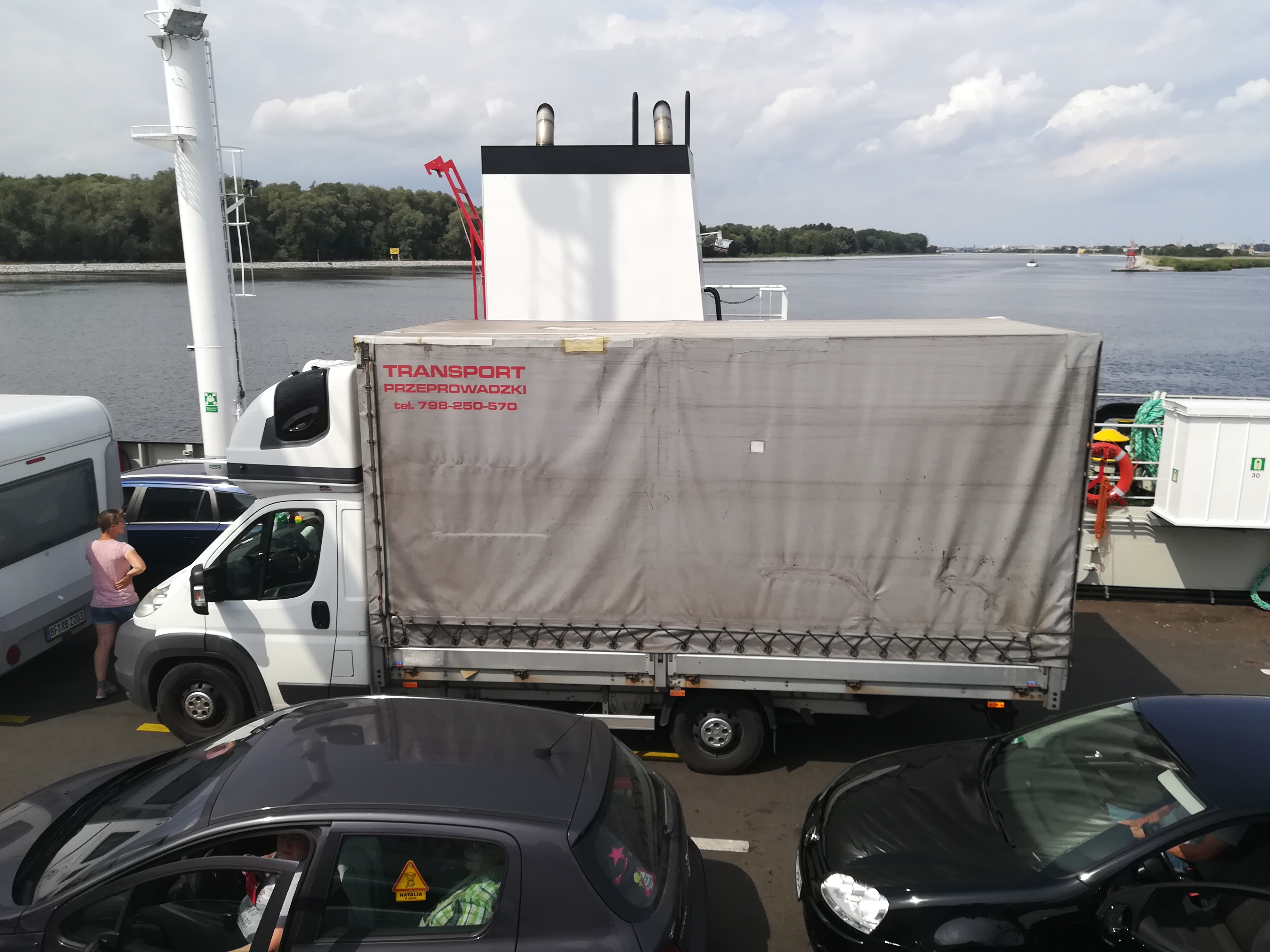 Biała ciężarówka z szarą plandeką z napisem 'TRANSPORT PRZEPROWADZKI tel. 798-250-570' na promie, w tle woda i drzewa.