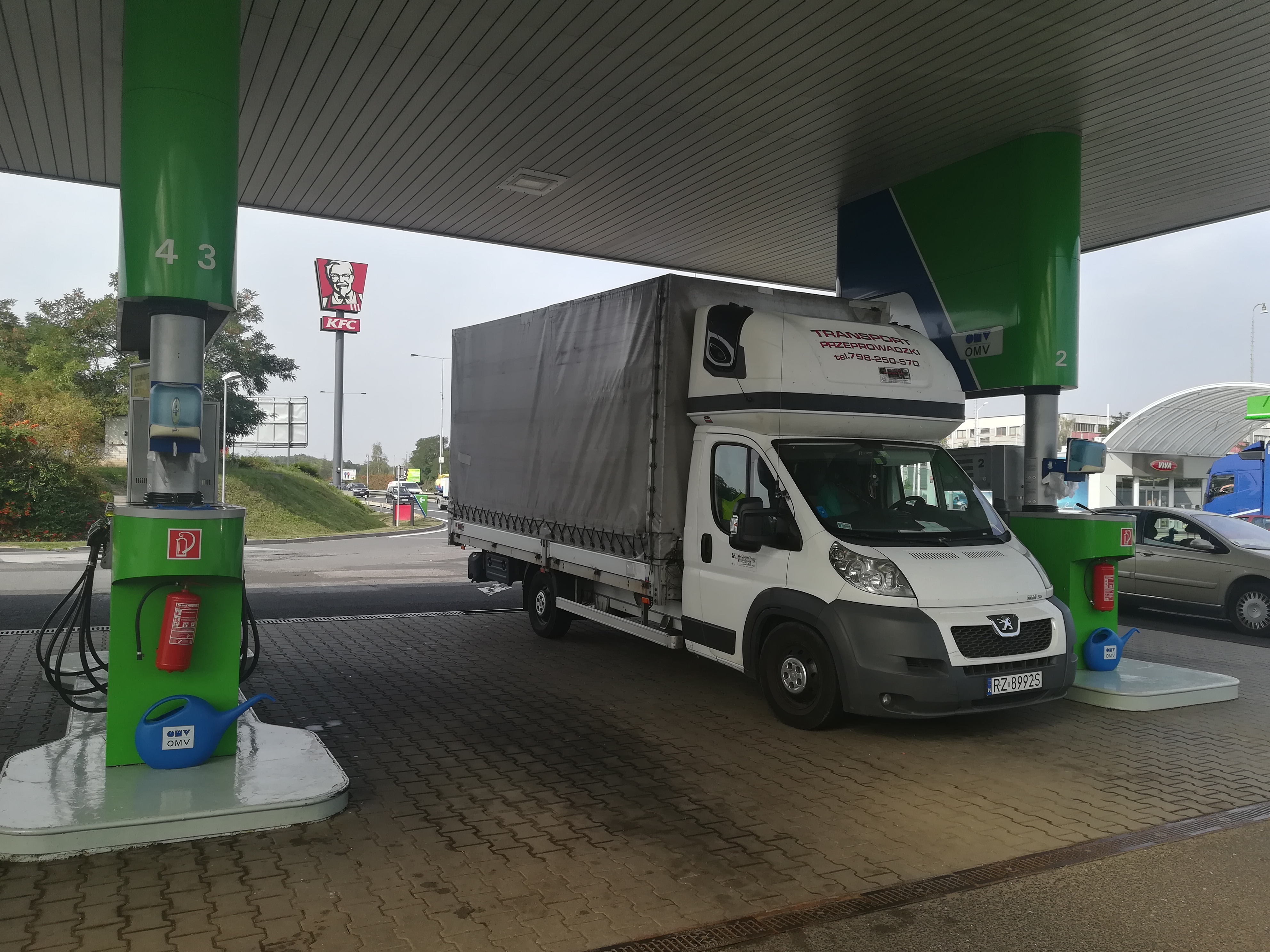 Biały samochód dostawczy z plandeką stoi na stacji benzynowej OMV, obok dystrybutora z niebieską konewką i czerwoną gaśnicą, w tle widoczny szyld KFC.