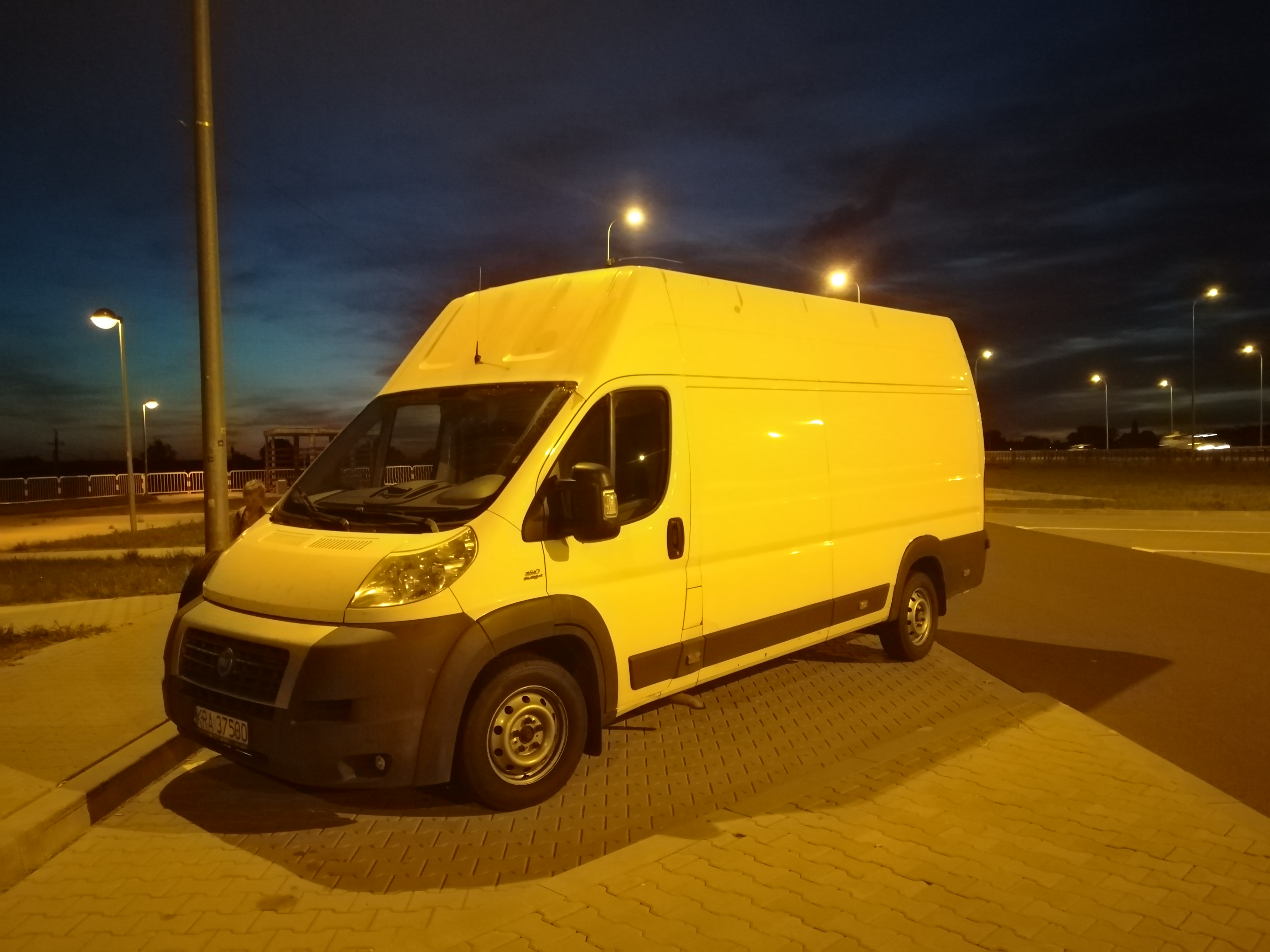 Biały bus dostawczy stoi zaparkowany na oświetlonym placu, widok z przodu na tle wieczornego nieba i lamp ulicznych.