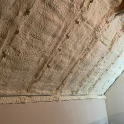 Poddasze ocieplone pianką poliuretanową, widoczne drewniane elementy konstrukcji dachu i biała, porowata struktura pianki. Ściana pomalowana na jasny kolor.