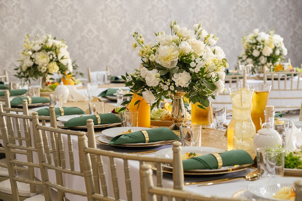 Elegancko nakryty stół z białym obrusem, złotymi podkładkami, zielonymi serwetkami ze złotymi pierścieniami, aranżacjami kwiatowymi w odcieniach bieli i kremu oraz napojami w szklanych dzbankach...