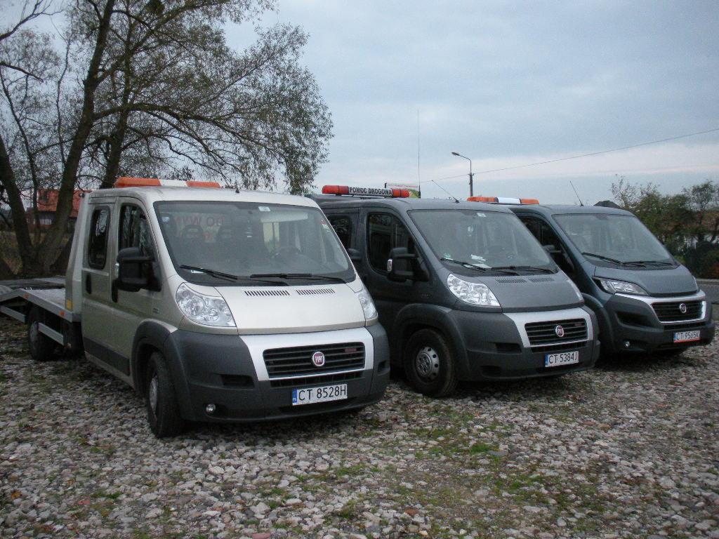 Trzy busy z platformami transportowymi zaparkowane na żwirowym podłożu, widoczne pomarańczowe światła ostrzegawcze na dachach.