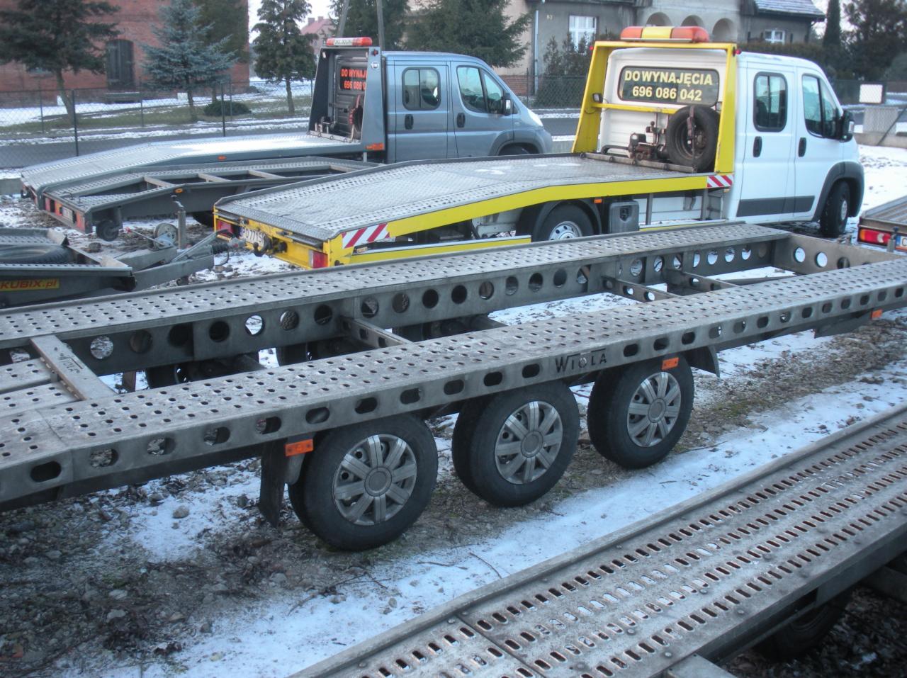 Szara, trzysiowa laweta samochodowa marki Wiola na śniegu, z lawetami i samochodami dostawczymi w tle.