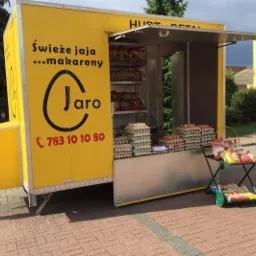 Żółty mobilny punkt sprzedaży z napisem 'Świeże ... makarony' i logo, oferujący kartony z produktami na ladzie i wewnątrz, dodatkowe produkty na wózku obok.