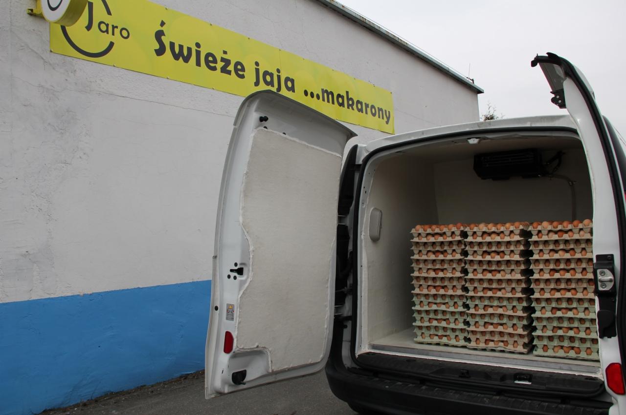 Transport świeżych produktów w chłodni samochodowej: otwarte drzwi furgonetki ukazują stosy wytłaczanek z produktami spożywczymi. Na ścianie budynku widoczny szyld reklamowy.