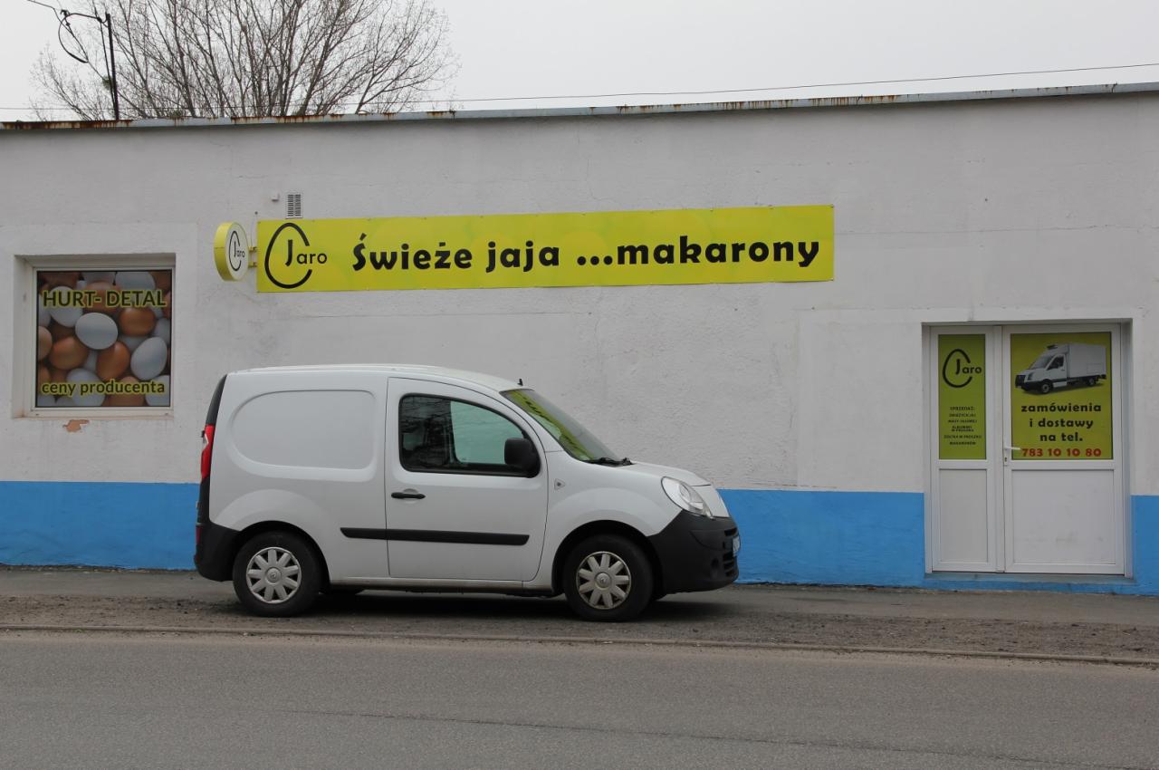 Biały samochód dostawczy zaparkowany przed budynkiem z szyldem 'Jaro Świeże jaja...makarony' i oknem z ekspozycją jaj kurzych.