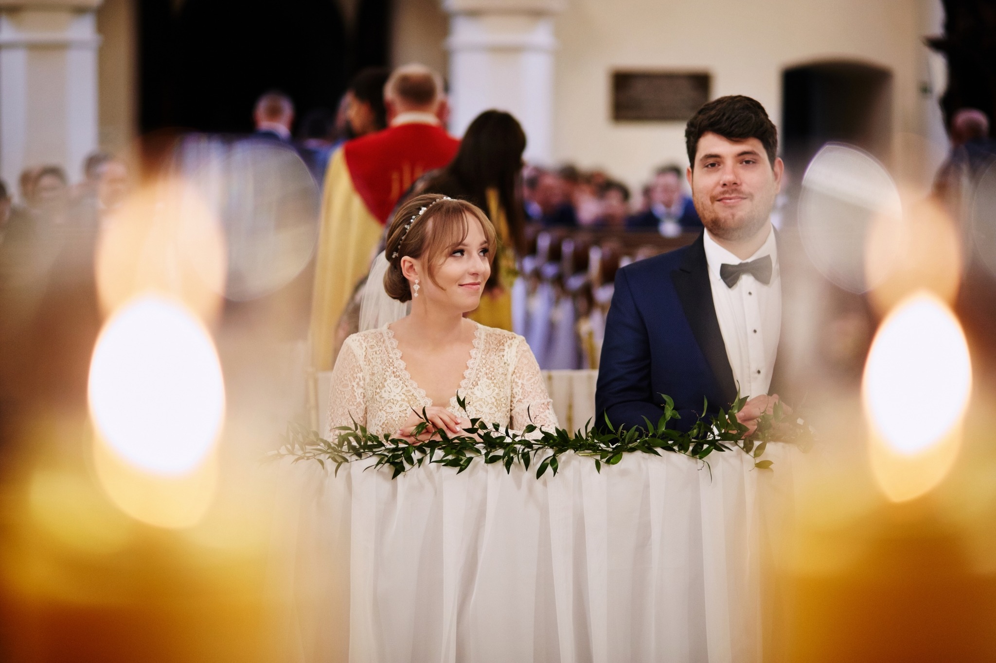 Para młoda siedząca za stołem udekorowanym zielonymi gałązkami podczas ceremonii ślubnej w kościele, z widocznymi gośćmi w tle.