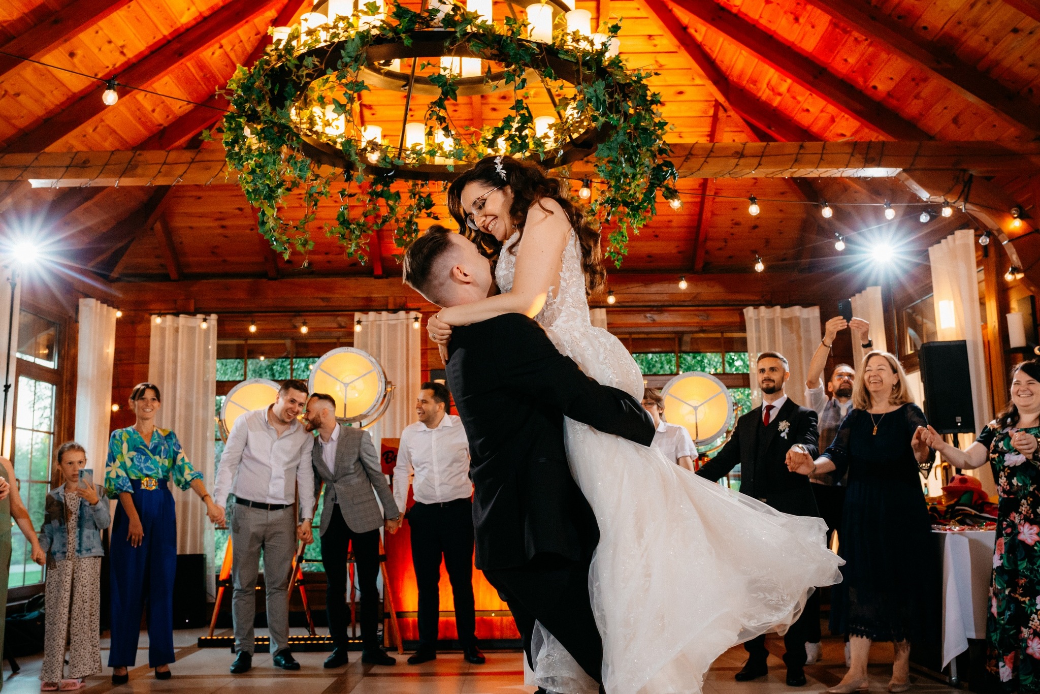 Uśmiechnięta para młoda w objęciach, panna młoda w białej sukni z koronką, pan młody w czarnym garniturze, taniec na parkiecie z drewnianą podłogą, goście weselni w tle, dekoracyjny żyrandol...