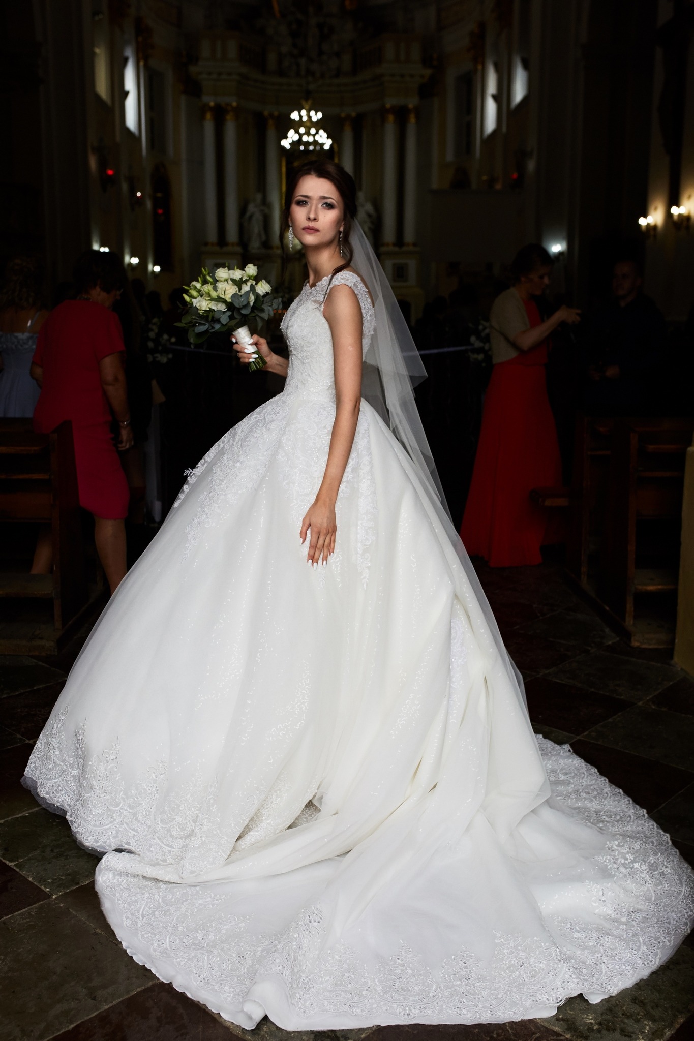 Panna młoda w bogato zdobionej sukni ślubnej z długim trenem i bukietem białych róż w kościele, w tle widoczni goście.