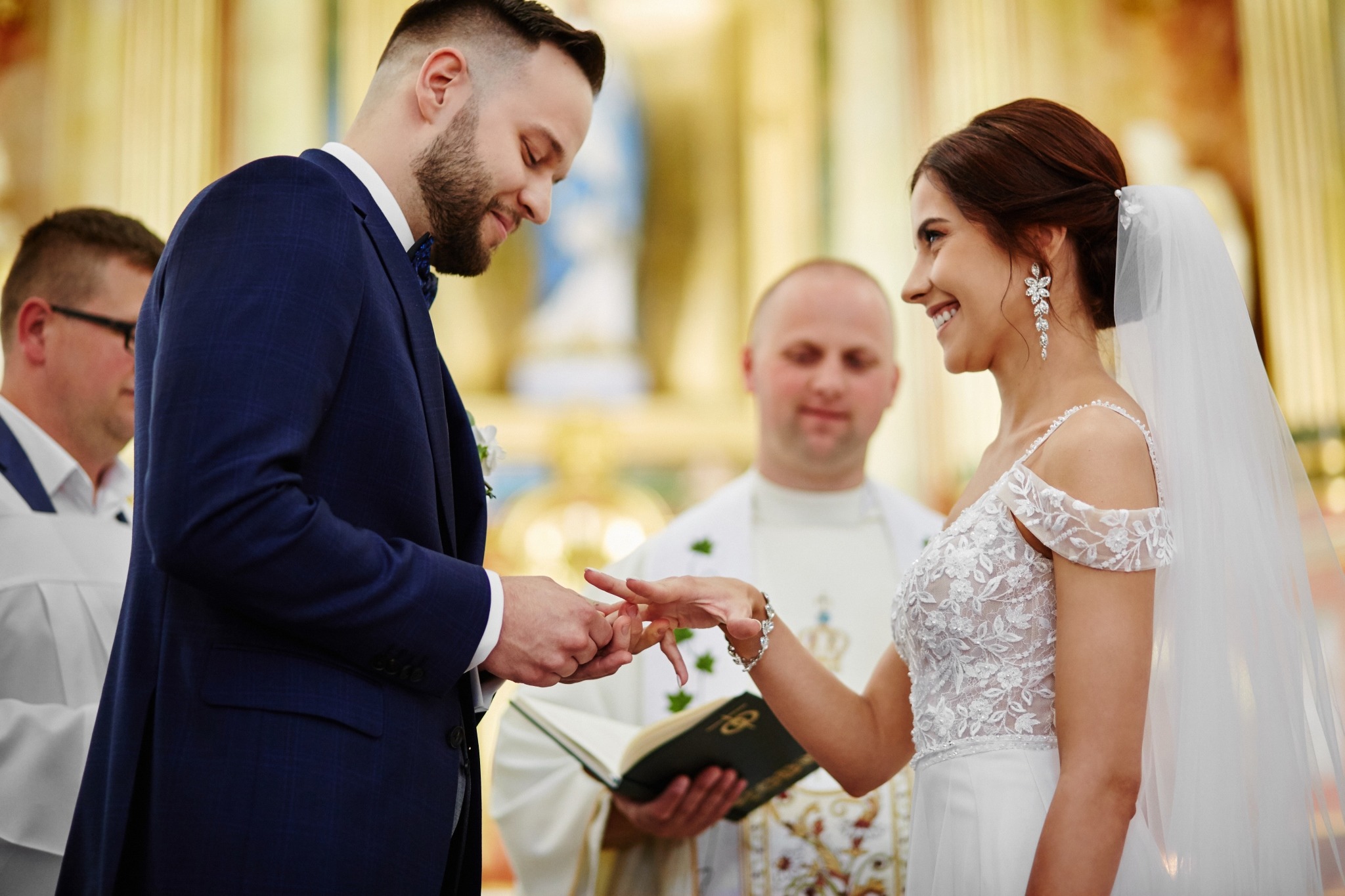 Moment zakładania obrączki podczas ceremonii ślubnej w kościele, panna młoda uśmiechnięta, w tle ksiądz z księgą.