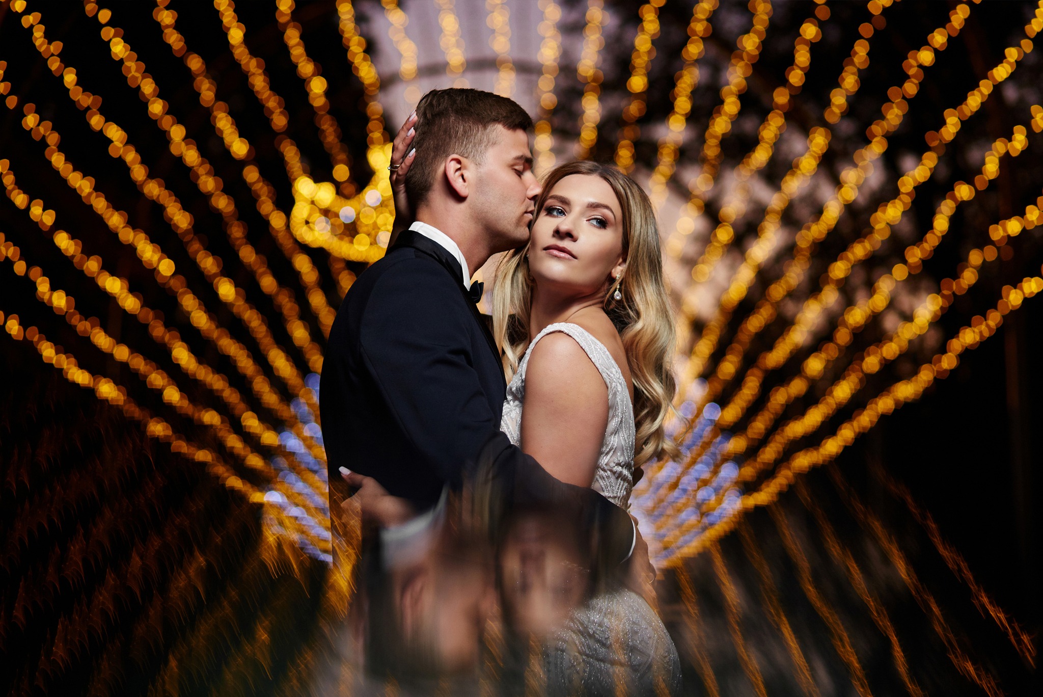 Elegancko ubrana para, mężczyzna w smokingu całuje kobietę w sukni ślubnej na tle rozmytych, dekoracyjnych światełek.