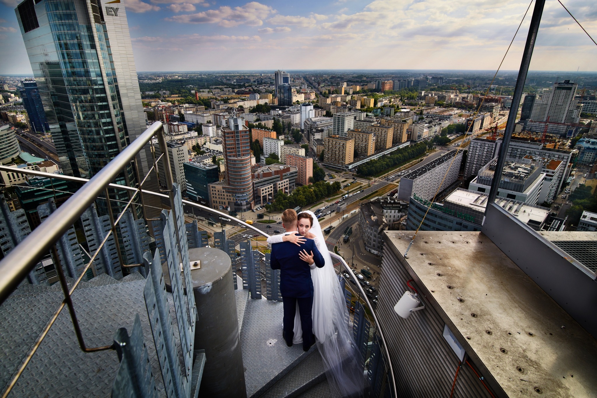Para młoda w objęciach na metalowych schodach wieżowca z panoramą miasta w tle, panna młoda spogląda w obiektyw.