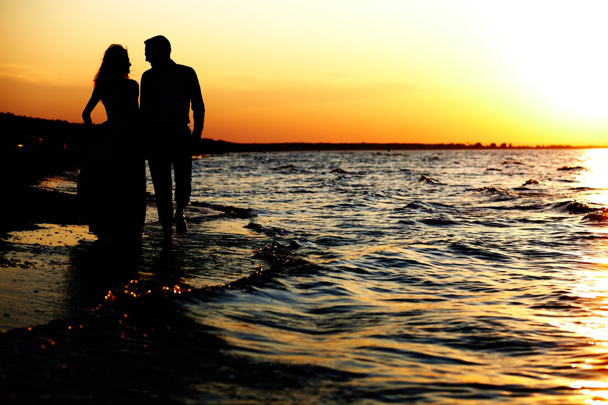Sylwetki pary spacerującej brzegiem morza o zachodzie słońca, ciepłe pomarańczowe światło odbija się w falach.