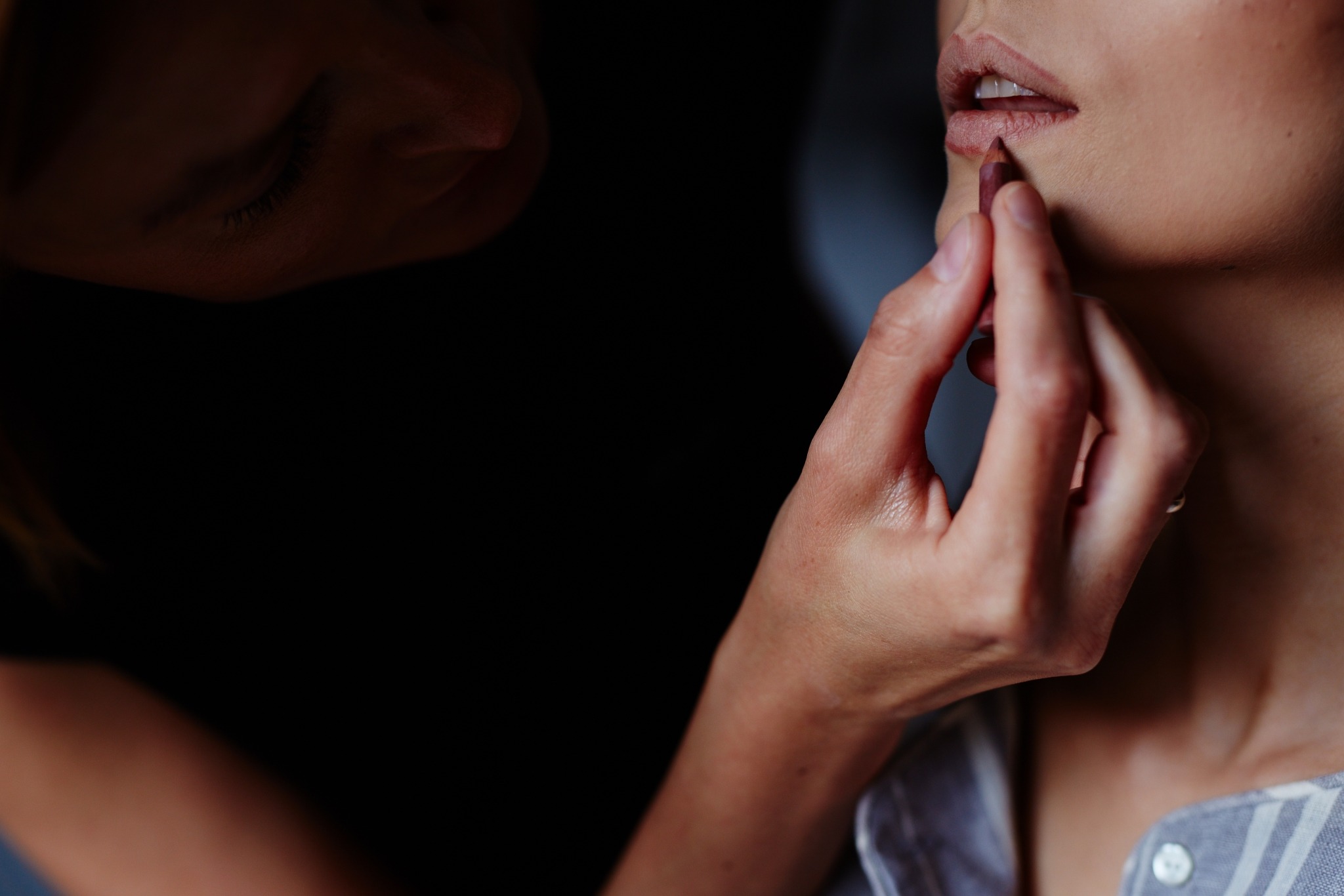 Makijażystka precyzyjnie obrysowuje usta modelki konturówką w odcieniu nude, zbliżenie na dłoń i twarz.