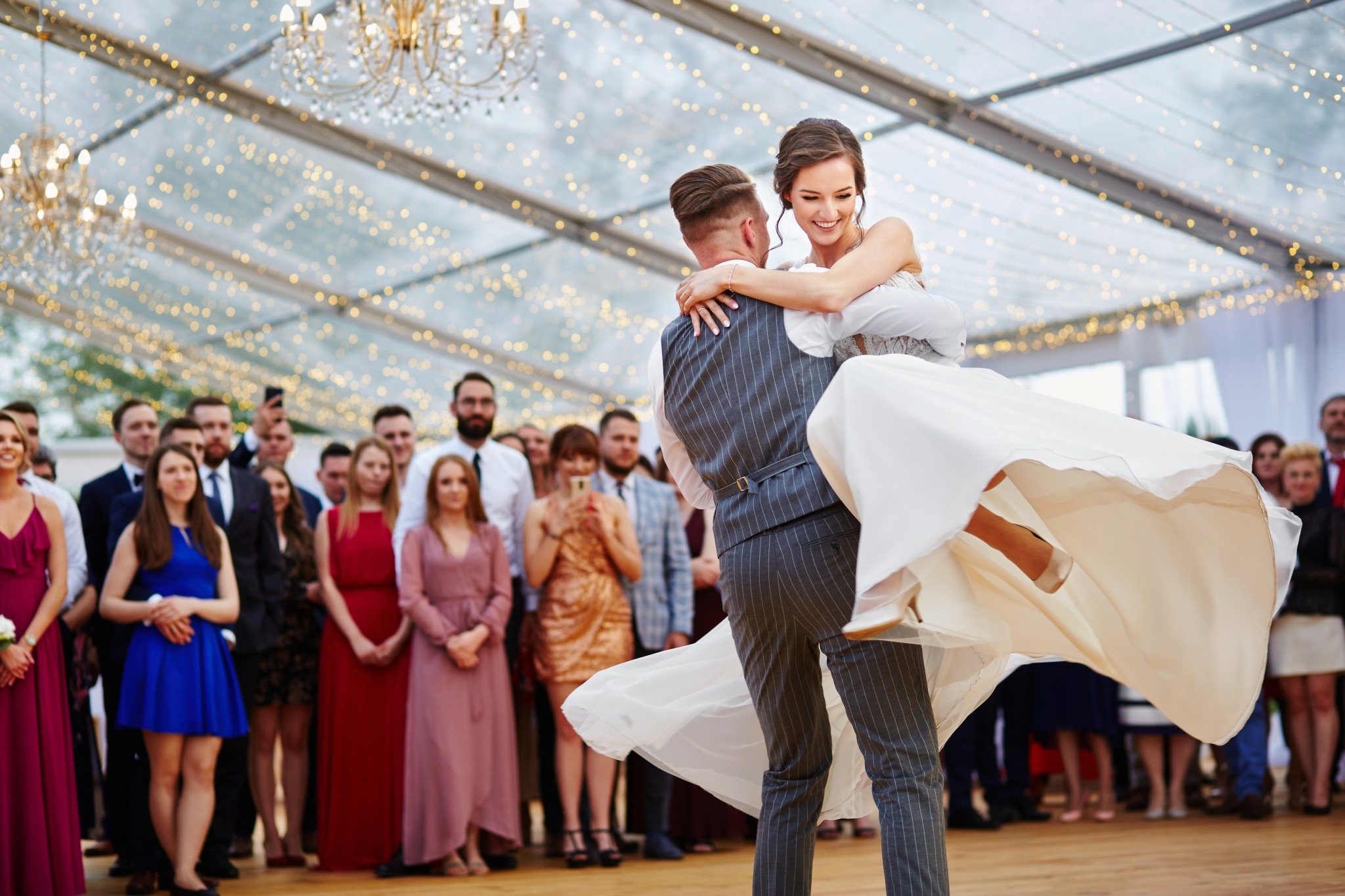 Uśmiechnięta panna młoda w białej sukni podnoszona przez pana młodego w szarym garniturze na tle gości weselnych i dekoracji świetlnych.