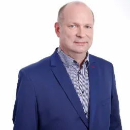 Portret mężczyzny w niebieskiej marynarce i wzorzystej koszuli, uśmiechającego się na białym tle.