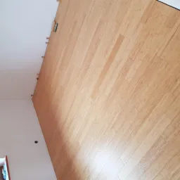 Ukośne ujęcie świeżo ułożonej podłogi z paneli w jasnym odcieniu drewna, widoczne listwy przypodłogowe i fragment ściany z gniazdkiem.