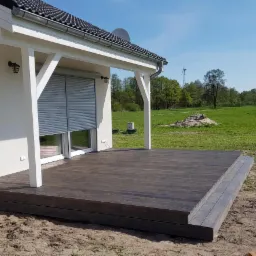 Nowoczesny taras z ciemnego drewna przy białym domu z roletami, widok na zielony trawnik i drzewa w tle, w oddali turbina wiatrowa.