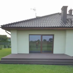 Nowoczesny taras kompozytowy z dwoma stopniami, przylegający do domu z brązowymi oknami i dachem z dachówki, otoczony zielonym trawnikiem.