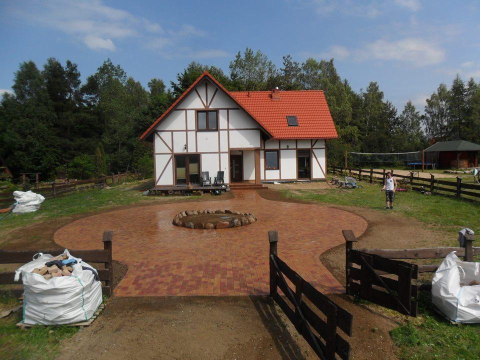 Ułożona kostka brukowa w kolorze czerwono-brązowym tworząca podjazd do domu z elementami dekoracyjnymi, widoczne worki z materiałem i fragment drewnianego ogrodzenia.