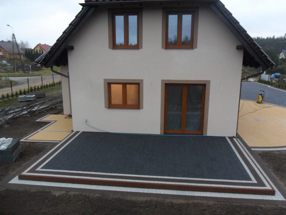 Wykończony taras z ciemnej kostki brukowej z dekoracyjnym obramowaniem z cegły i jasnych płytek, przylegający do ściany domu z drewnianą stolarką okienną i drzwiową, widoczny fragment kostki...