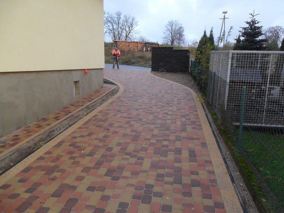 Zakrzywiony podjazd z kostki brukowej w odcieniach czerwieni i brązu, obrzeża z drobnego kruszywa, widoczny fragment budynku z pomarańczową piłką przy ścianie i osoba w odblaskowej kamizelce w oddali.