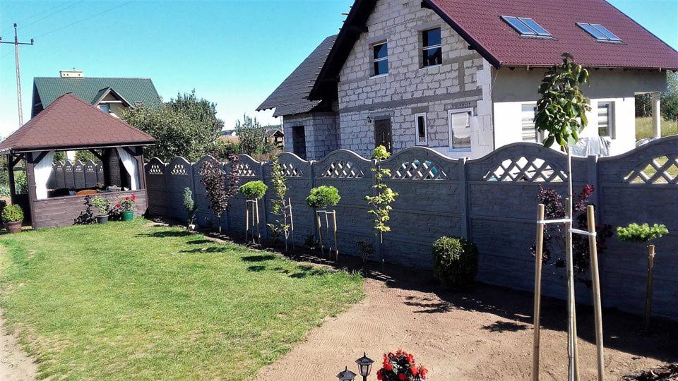 Prefabrykowane betonowe ogrodzenie z dekoracyjnymi wycięciami w kształcie rombów, oddzielające trawnik od świeżo wyrównanej ziemi, z młodymi drzewkami podpartymi palikami i budynkiem w stanie...