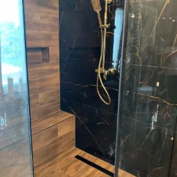 Nowoczesna kabina prysznicowa z czarnymi płytkami imitującymi marmur z złotymi żyłami, złotą armaturą i drewnopodobnymi płytkami na podłodze i obudowie wnęki, widoczny odpływ liniowy.