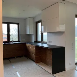 Nowoczesna kuchnia z drewnianymi szafkami dolnymi i białymi górnymi, czarny blat, widok na okno i drzwi balkonowe, podłoga w trakcie wykończenia.