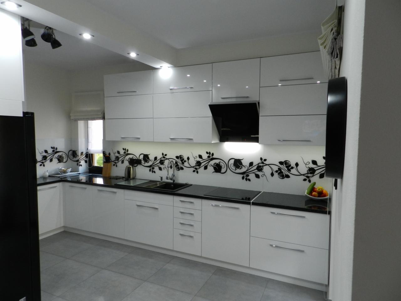 Nowoczesna, biała kuchnia z czarnym blatem i dekoracyjną naklejką z motywem roślinnym na ścianie między szafkami, oświetlenie punktowe na suficie.