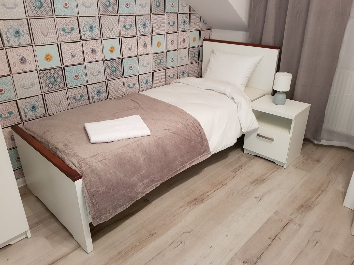 Pokój z pojedynczym łóżkiem, białą szafką nocną i lampką, na ścianie tapeta w geometryczne wzory imitujące szuflady.