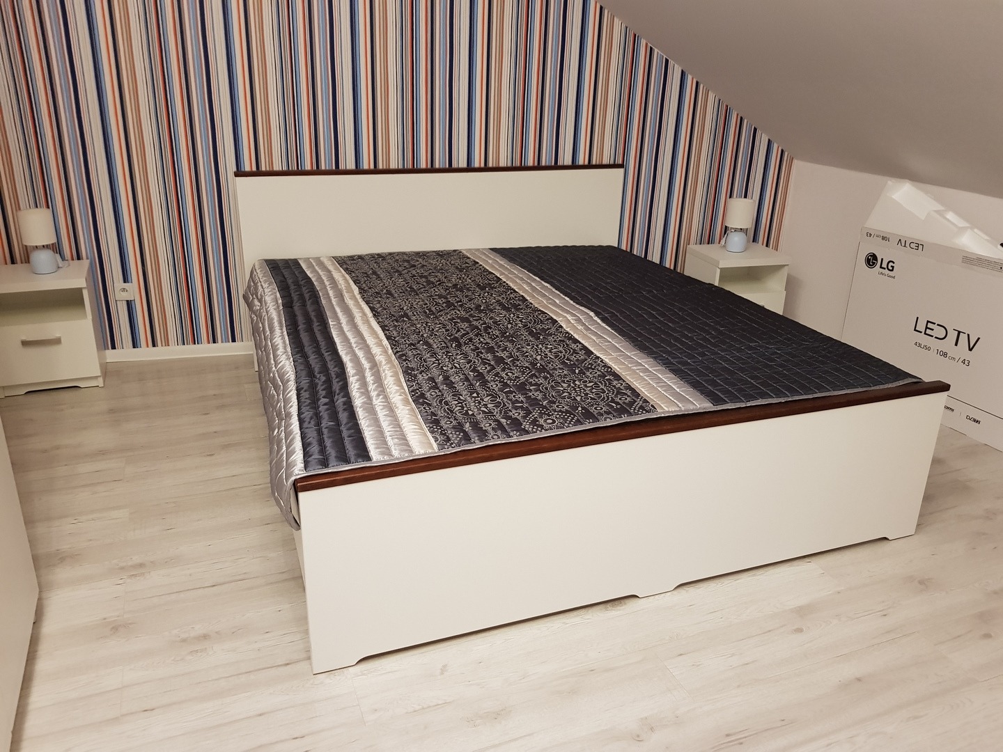 Białe łóżko z ciemnobrązowym obramowaniem, nakryte wzorzystą kołdrą w odcieniach szarości i srebra, w pokoju z tapetą w pionowe pasy i szafkami nocnymi z lampkami.