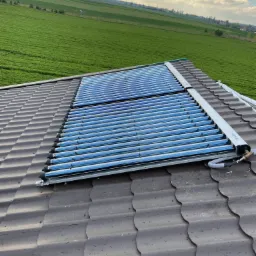 Kolektory słoneczne rurowe zamontowane na brązowym dachu z widokiem na zielone pole i zachmurzone niebo.