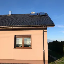 Dom jednorodzinny z panelami słonecznymi na dachu i nową rynną spustową. Widoczne dwa okna z roletami zewnętrznymi i firankami wewnątrz.