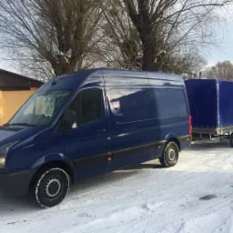 Niebieski bus z przyczepą plandekową na śniegu w zimowy dzień, widoczne ślady opon, budynek w tle.