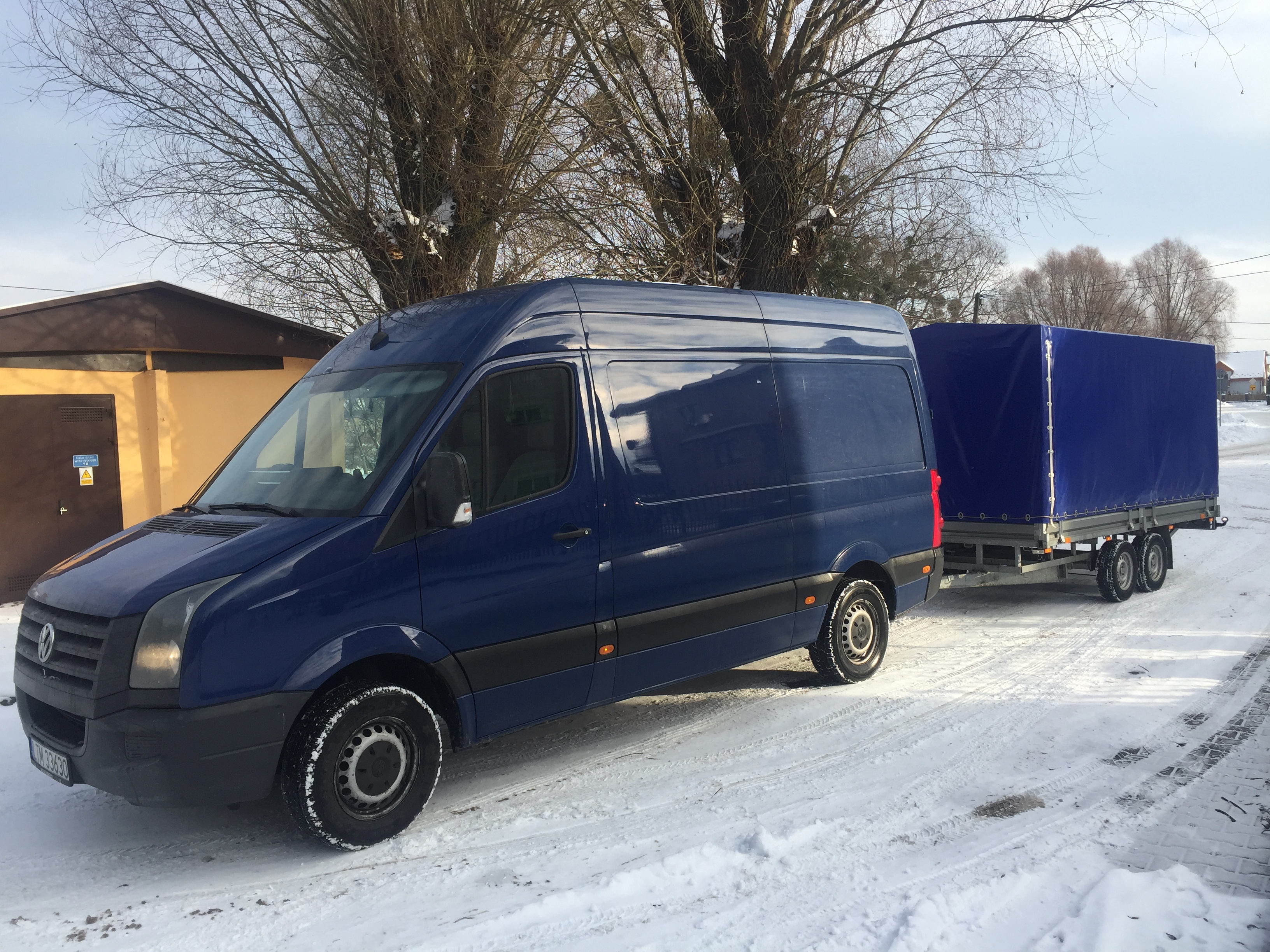 Niebieski bus z przyczepą plandekową na śniegu w zimowy dzień, widoczne ślady opon, budynek w tle.