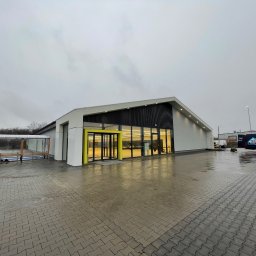 House Investments Piotr Ziółkowski - Nowoczesny budynek usługowy z elementami elewacji w kolorze białym i czarnym, żółte obramowanie wejścia, widoczne zaparkowane busy dostawcze na zewnątrz, pochmurny dzień.
