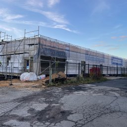 House Investments Piotr Ziółkowski - Budynek w trakcie ocieplania, widoczne rusztowania, materiały izolacyjne oraz pracownicy w odblaskowych kamizelkach. Baner reklamowy na siatce ochronnej.