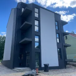 Nowoczesny budynek wielorodzinny z balkonami, w trakcie prac wykończeniowych elewacji. Widoczne rusztowania i materiały budowlane na terenie posesji.