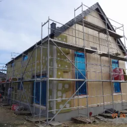 Dom jednorodzinny w trakcie ocieplania, widoczne rusztowania, izolacja termiczna z wełny mineralnej oraz zabezpieczone okna folią. Częściowo ocieplony fragment ściany z widocznymi łatami.