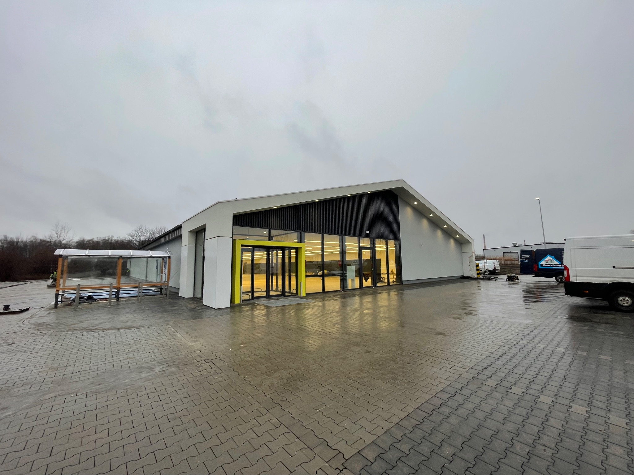 Nowoczesny budynek usługowy z elementami elewacji w kolorze białym i czarnym, żółte obramowanie wejścia, widoczne zaparkowane busy dostawcze na zewnątrz, pochmurny dzień.