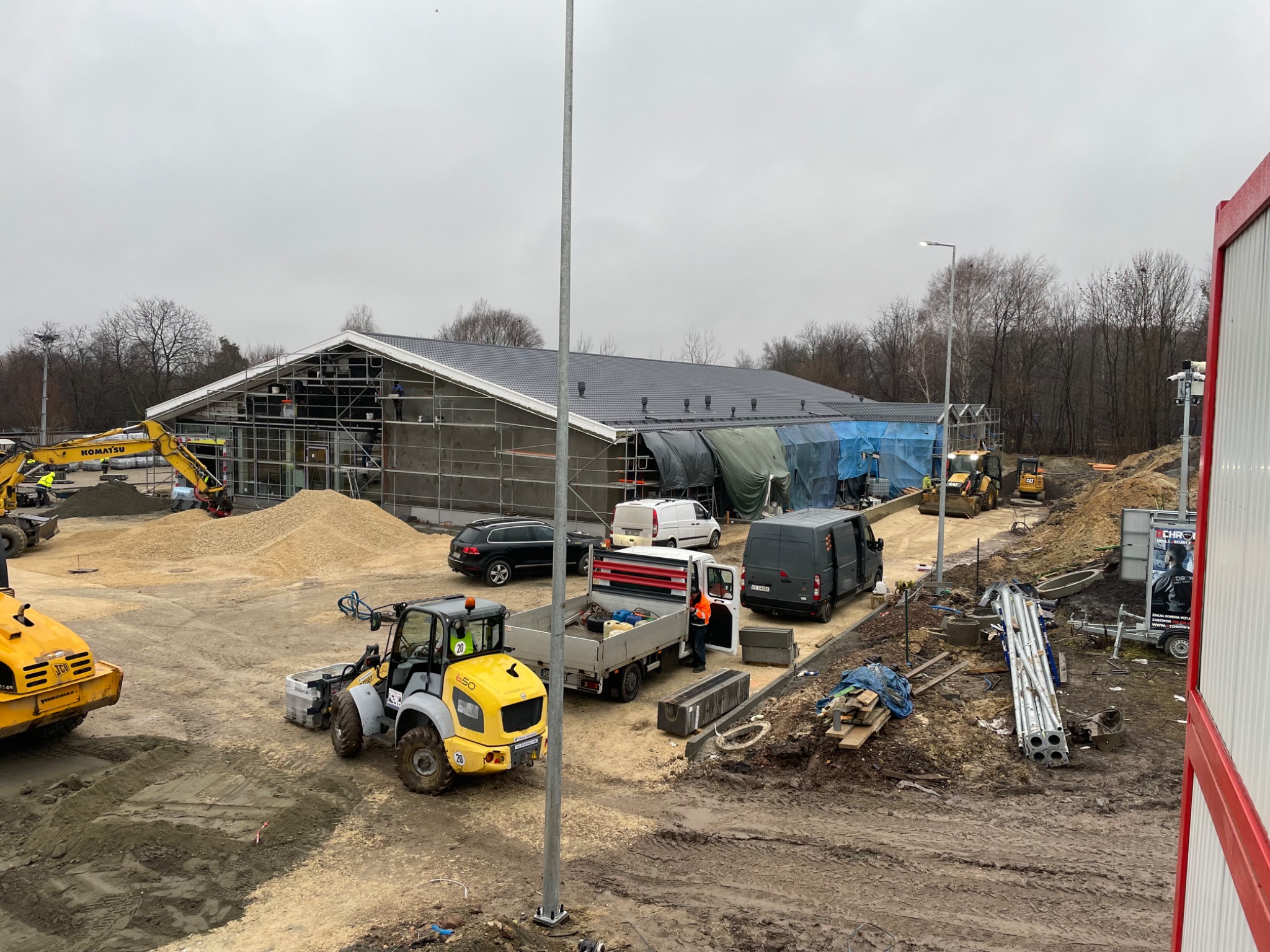 Budynek w trakcie prac elewacyjnych, widoczne rusztowania, maszyny budowlane i materiały, pochmurne niebo.