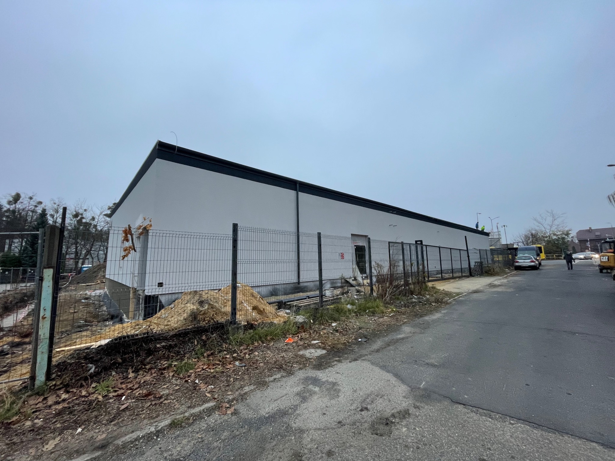 Budynek w trakcie prac elewacyjnych, widoczne rusztowania i materiały budowlane, teren częściowo ogrodzony, pochmurne niebo.