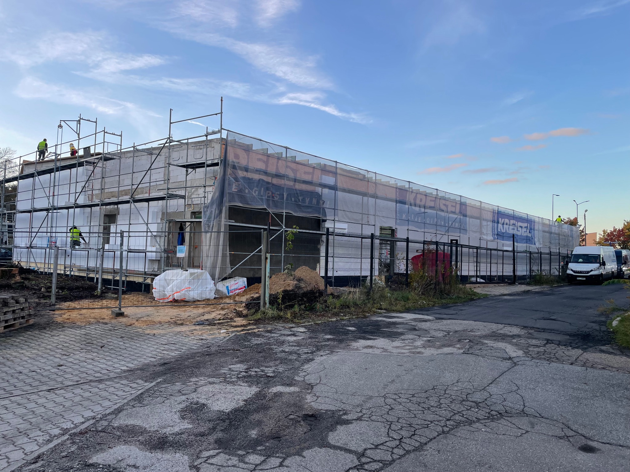 Budynek w trakcie ocieplania, widoczne rusztowania, materiały izolacyjne oraz pracownicy w odblaskowych kamizelkach. Baner reklamowy na siatce ochronnej.