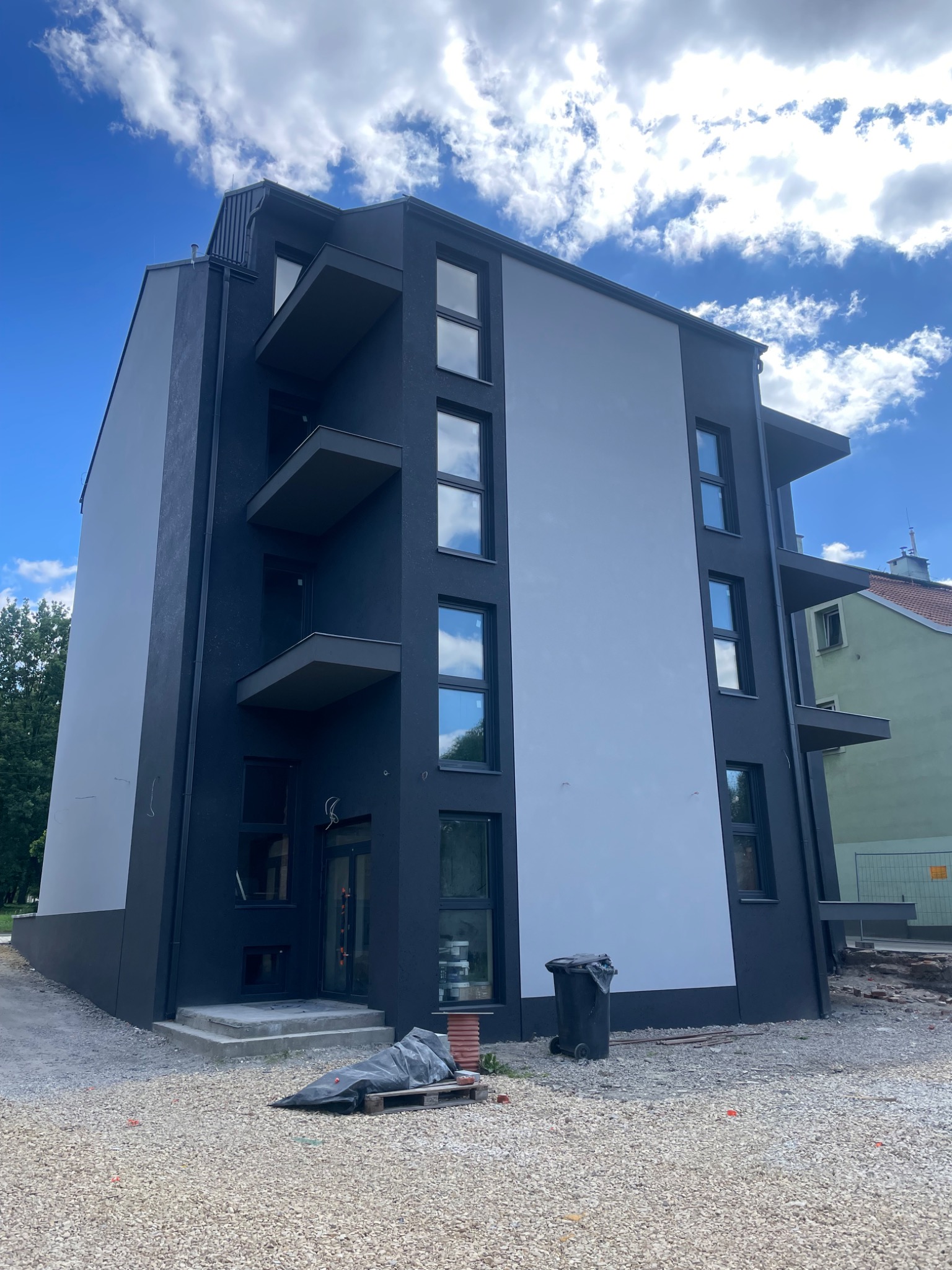 Nowoczesny budynek wielorodzinny z balkonami, w trakcie prac wykończeniowych elewacji. Widoczne rusztowania i materiały budowlane na terenie posesji.